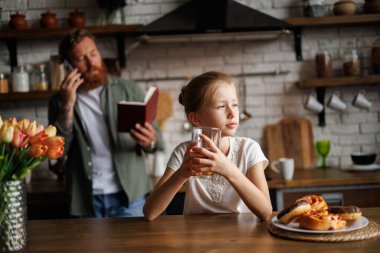 Tatlı pastanın yanında meyve suyu tutan sıkkın ergen kız ve mutfakta not defteriyle meşgul ebeveyn. 