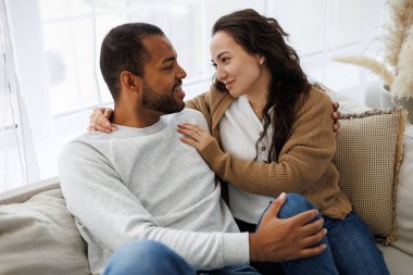 Gülümseyen genç Asyalı kadın, evde kanepede dinlenirken Afrikalı Amerikalı erkek arkadaşına sarılıyor.