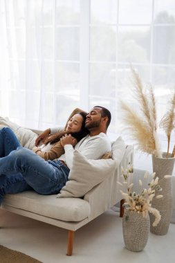 Gülümseyen Afrikalı Amerikalı genç adam evinde kanepede otururken rahat Asyalı kız arkadaşına sarılıyor.