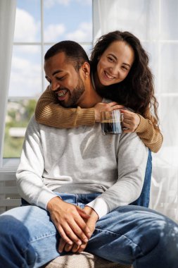 Gülümseyen Asyalı bir kadın kahve tutuyor ve sabah evde Afrikalı Amerikalı erkek arkadaşına sarılıyor.