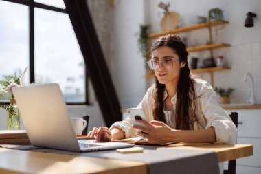 Evde çevrimiçi kurslar sırasında bilgisayar ve akıllı telefon kullanan gözlüklü kadın.