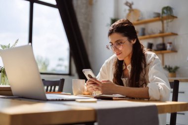 Gözlüklü gülümseyen kadın evde sabahları kitapların ve laptopun yanında akıllı telefon kullanıyor.