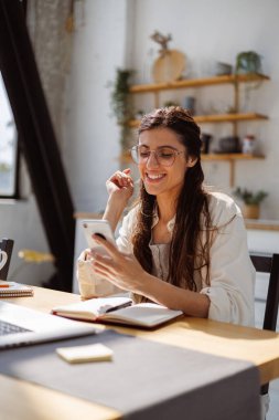 Gözlüklü neşeli kadın dizüstü bilgisayarın yanında akıllı telefon ve mutfaktaki masanın üstünde not defteri kullanıyor. 