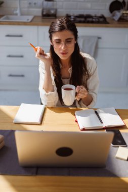 Gözlüklü kadın evde online eğitim sırasında dizüstü bilgisayar ve not defterlerinin yanında kalem ve bardak tutuyor.