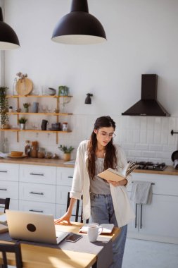 Cihazların yanında kitap okuyan kadın, evdeki çevrimiçi kurslarda masanın üzerinde fincan okuyor.