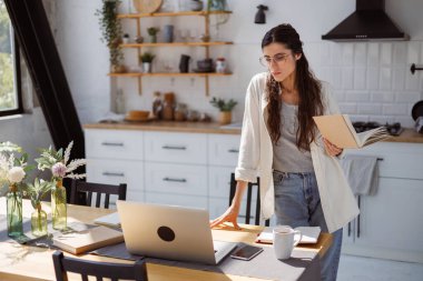 Kitapları tutan ve mutfaktaki fincanın yanında dizüstü bilgisayara bakan kadın, kendini geliştirme konsepti.