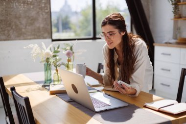 Gözlüklü, gülümseyen, elinde akıllı telefon ve bardakla sabahları mutfakta dizüstü bilgisayarın yanında gülümseyen. 
