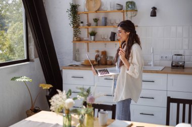 Gözlüklü gülümseyen kadın çevrimiçi eğitim sırasında video görüşmesi yapıyor. Mutfakta dizüstü bilgisayarda. 