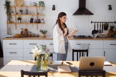 Gözlüklü bir kadın mutfakta aletlerin yanında kitap okuyor, uzaktan öğrenme konsepti.