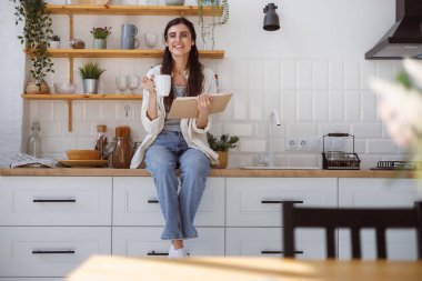 Gözlüklü mutlu kadın kameraya bakıyor ve modern mutfakta kitap ve bardak tutuyor. 