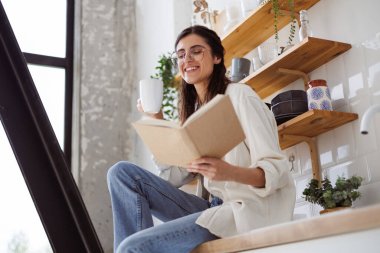 Gözlüklü, elinde bardak olan ve modern mutfakta kitap okuyan pozitif bir kadın.