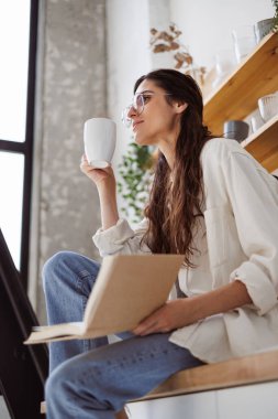 Gözlüklü rahat bir kadın sabah mutfakta bardak ve kitap tutuyor. 