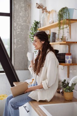 Modern mutfakta atölyede otururken elinde bardak ve kitap tutan pozitif bir kadın. 