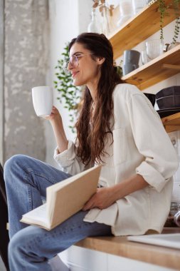 Gözlüklü gülümseyen kadın, elinde bardak ve mutfakta bulanık bir kitap tutuyor. 
