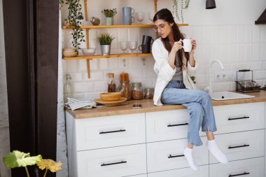 Gözlüklü bir kadın elinde bardak ve mutfakta siyah ekranlı akıllı bir telefon tutuyor.