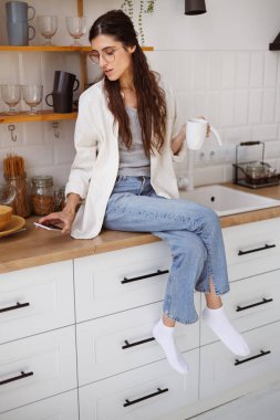 Gözlüklü bir kadın elinde fincanla mutfakta otururken akıllı telefonuna bakıyor.