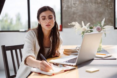 Evdeki cihazların yanındaki deftere kablosuz kulaklık takan serbest çalışan bir kadın.