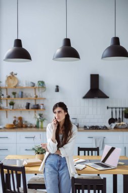 Ciddi kadın serbest çalışan, elinde gözlük olan, akıllı telefonla konuşan, dizüstü bilgisayarın yanında duran ve mutfakta not defterleri olan biri.