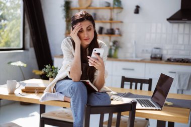 Sinirli kadın serbest çalışan bilgisayarın yanında akıllı telefon ve mutfakta not defteri kullanıyor. 