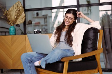 Gözlüklü, gülümseyen, evinde koltukta otururken dizüstü bilgisayar kullanan serbest çalışan bir kadın.