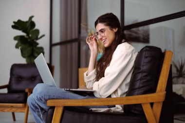 Gözlüklü, gülümseyen, evinde rahat koltukta dizüstü bilgisayar kullanan serbest çalışan bir kadın.