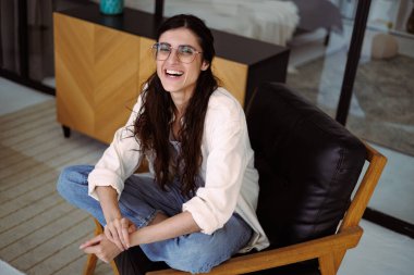 Gözlüklü, sıradan giysileri olan neşeli esmer kadın evdeki koltukta otururken kameraya bakıyor.