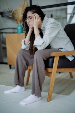 Gözlüklü depresif kadın evde koltukta otururken duygusal acıdan muzdarip.