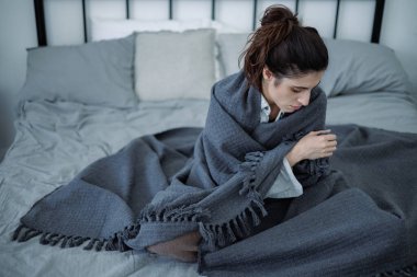 Evde yatakta otururken battaniyeye sarılmış depresif esmer kadın, yalnızlık konsepti.