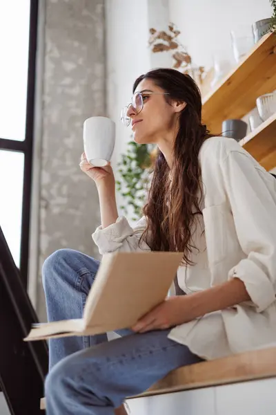 Gözlüklü rahat bir kadın sabah mutfakta bardak ve kitap tutuyor. 