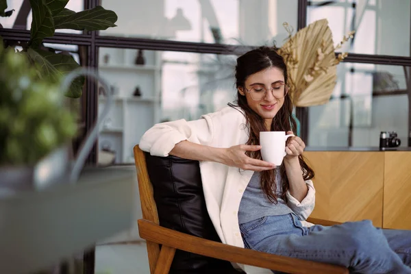 Gözlüklü, gülümseyen esmer kadın modern evdeki koltukta dinlenirken elinde bardak tutuyordu.