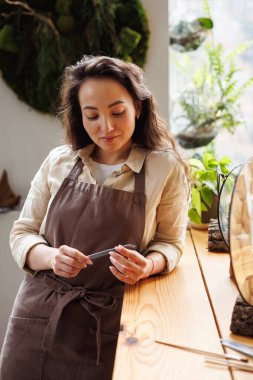 Önlüklü genç Asyalı zanaatkar çiçekçide çerçevelenmiş otların yanında cımbız tutuyor. 