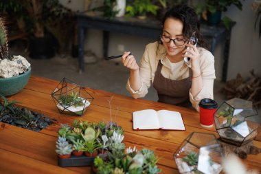 Gülümseyen Asyalı çiçekçi defterin, çiçekçilerin ve çiçekçinin yanındaki akıllı telefondan konuşuyor.