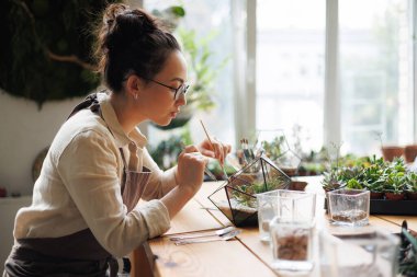 Asyalı çiçekçinin bahçe aletleriyle profili çiçek dükkanındaki sulu çiçeklerin yanındaki teraryumda küçük bir bahçe yaratıyor.