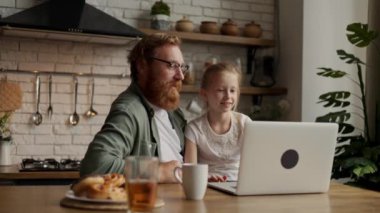 Gülümseyen baba ve kız konuşuyor ve bilgisayardan mutfakta kahvaltıya yakın bir yerde video görüşmesi yapıyorlar.