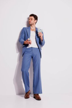 Mavi takım elbiseli ve kolsuz tişörtlü şık bir adam gri duvarın yanında dolar tutuyor. 