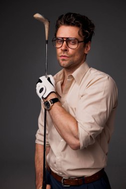 Gözlüklü ve eldiven takan iyi giyimli bir adam gri alanda golf sopası tutuyor. 