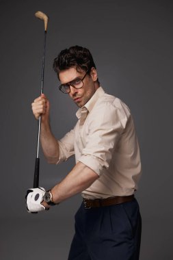 Klasik kıyafetli ve eldivenli esmer adam golf sopası tutuyor ve gri renkli kameraya bakıyor. 