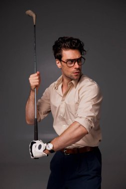 Gözlüklü ve eldivenli modaya uygun bir adam golf sopası tutuyor ve yüzünü griye çeviriyor. 