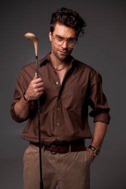 Kahverengi gömlekli, gözlüklü, golf sopası tutan esmer bir adam. 