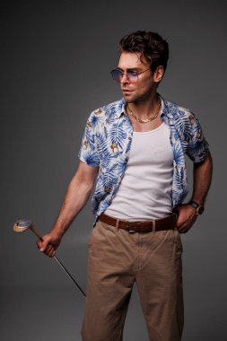 90 'ların kıyafetleri ve güneş gözlüklü şık bir adam. Golf sopası gri alanda izole edilmiş. 