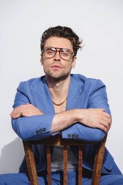 Mavi takım elbiseli, gözlüklü, gri sandalyede oturan şık bir adam.