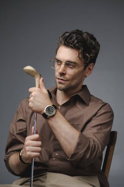 Klasik kıyafetli esmer adam ve lüks gözlüklü golf sopasını gri sandalyede tek başına tutan adam. 