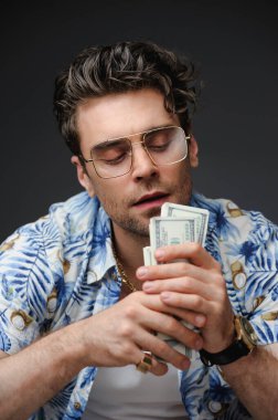 Aloha gömlekli ve gözlüklü iyi giyimli adamın portresi gri üzerinde izole edilmiş nakit para taşıyor.
