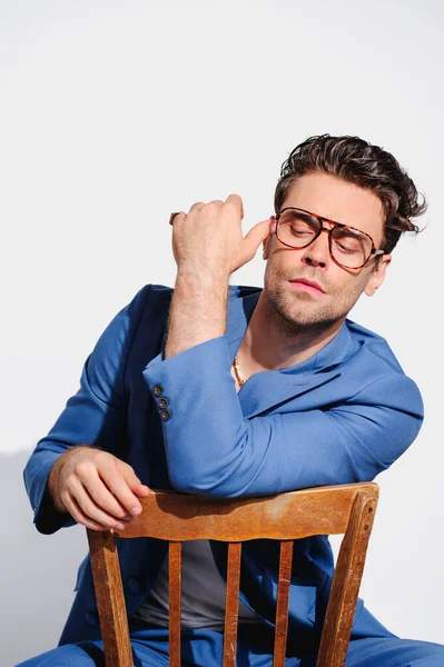 Mavi ceketli, altın zincirli ve gözlüklü, gri sandalyede poz veren şık bir adam. 