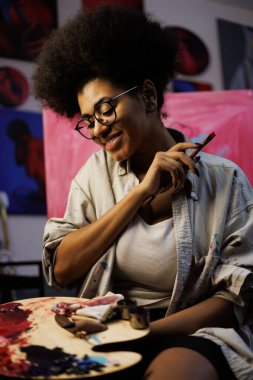 Neşeli Afrikalı Amerikalı kadın sanatçı, resim stüdyosunda boya paleti ve fırça tutuyor.