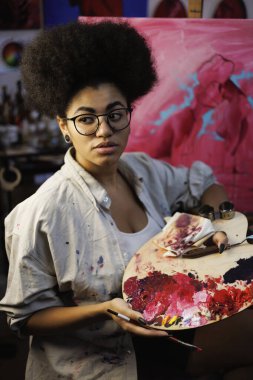 Afrikalı Amerikalı sanatçı atölyede bulanık tuvalin yanında boya paleti ve sanat malzemeleri tutuyor.