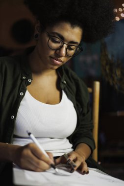 Gözlüklü Afrikalı Amerikalı ressamın çizim defterine ışıklandırmalı bir resim çizmesi. 