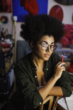 Kalem tutan Afrikalı Amerikalı sanatçı stüdyodaki bulanık resimlerin yakınındaki kameraya bakıyor.