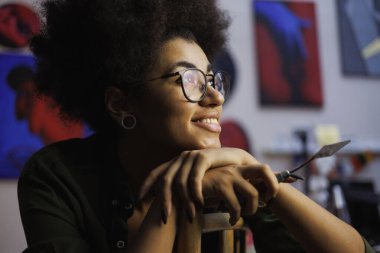 Gözlüklü gülümseyen Afrikalı Amerikalı ressamın portresi. Bulanık stüdyoda resim bıçağı tutuyor.