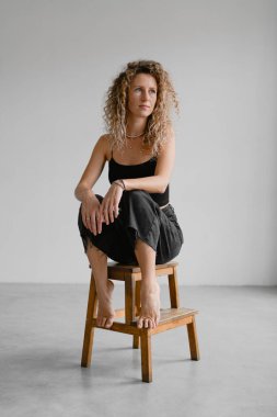 Koyu renk kotlu çıplak ayaklı kadın ve üstü başı gri arka planda tahta taburede oturan.